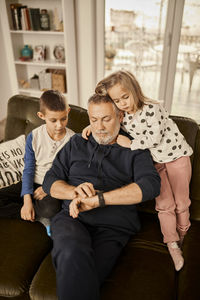 Boy and girl sitting with grandfather using smart watch sitting on sofa at home