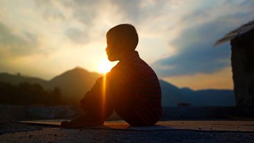 Side view of silhouette boy sitting on road against sky during sunset