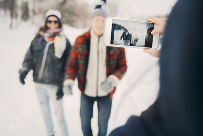 Woman photographing friends through smart phone on field during winter