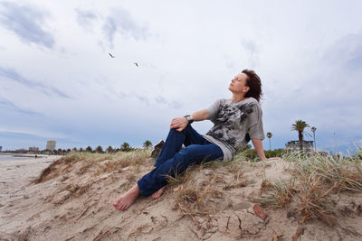 Full length of young woman sitting on beach against sky