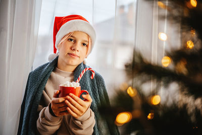 Young teen girl in santa hat having a cup of hot chocolate with marshmallow with christmas tree on