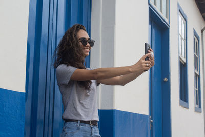 Smiling young woman taking selfie while standing by wall