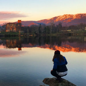 Rear view of man sitting by lake against sky during sunset