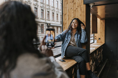 Mature female owner photographing young colleague sitting with hand in hair on window sill