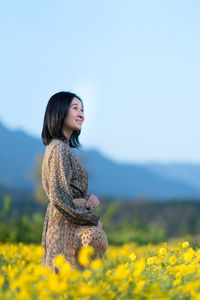 Young woman standing by yellow flower on field against sky