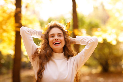 Portrait of smiling young woman with arms raised standing against trees