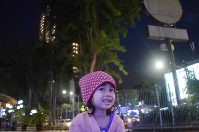 Smiling girl looking away while sitting against illuminated building at night