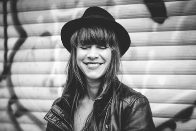 Smiling woman with eyes closed wearing hat against graffiti shutter in city