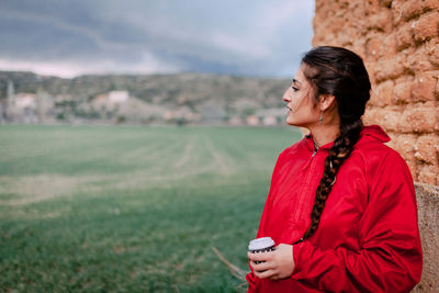 Young woman looking away holding drink standing by wall