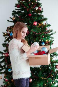 Teenage girl looking at christmas ornament while sitting by christmas tree at home