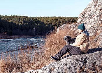 Young woman sitting on stone hugging dog looking at river  with first ice in fall or winter, beige 