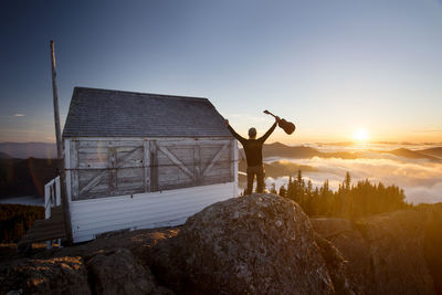 Rear view of man holding guitar while standing by log cabin on mountain during sunset