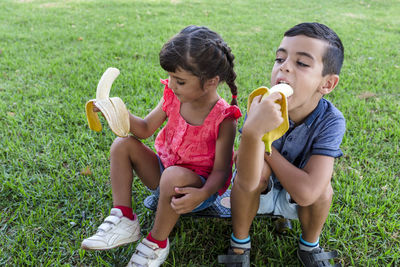 Two kids eating banana sitting outdoors on the grass in a park.