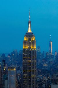 Illuminated empire state building amidst towers in city against blue sky