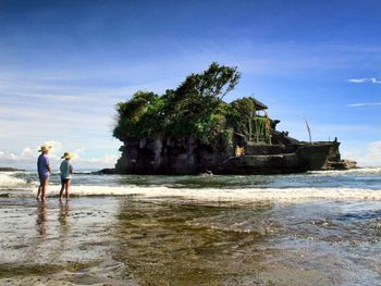 Rear view of sibling standing on shore at tanah lot against sky