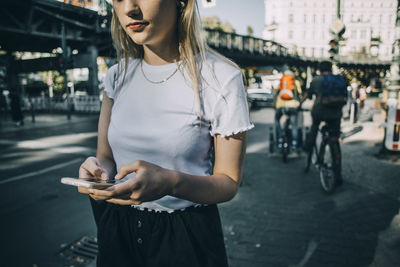 Midsection of young woman using smart phone while standing on sidewalk in city