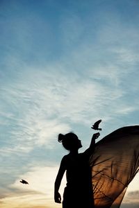 Silhouette woman reaching bird against sky during sunset