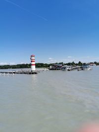 Lighthouse by sea against buildings against clear blue sky