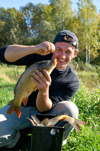 Portrait of smiling mature man holding fish while crouching on grassy field