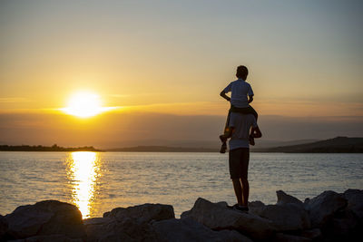 Rear view of father carrying son on shoulders while standing at beach against sky during sunset