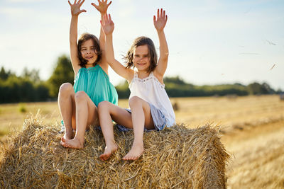 Two charming girls are sitting on a roll of mown rye in a field in summer