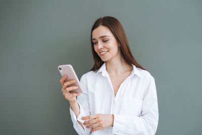 Portrait of smiling woman with dark long hair in white shirt using mobile phone on grey background 