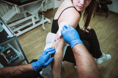 Back view crop anonymous male tattooist in latex gloves wiping freshly made tattoo on arm of female client with napkin while working in salon