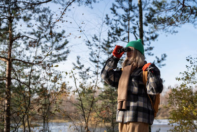 Middle aged woman tourist birdwatcher in autumn forest looking through binoculars on birds.