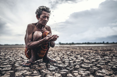 Shirtless man holding falling water while crouching on cracked land against sky