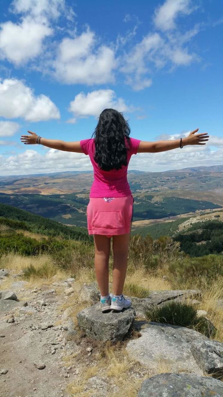 rear view, arms raised, real people, arms outstretched, sky, leisure activity, day, one person, cloud - sky, outdoors, pink color, nature, rock - object, lifestyles, beauty in nature, mountain, full length, scenics, standing, human body part, people