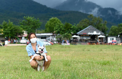 A woman wearing a mask hug a white french bulldog on the lawn looking at the camera.