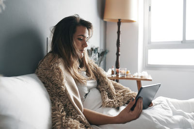 Sick young woman using digital tablet on bed against wall at home