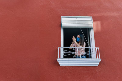 Young brunette woman with mask clapping from the window of the house with red background