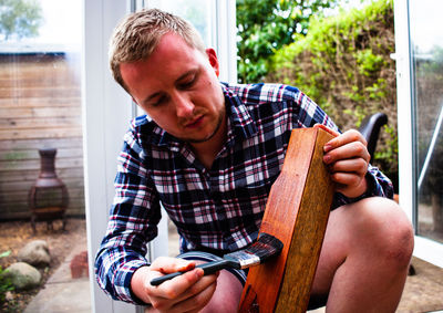 Young man painting wooden plank while crouching in yard