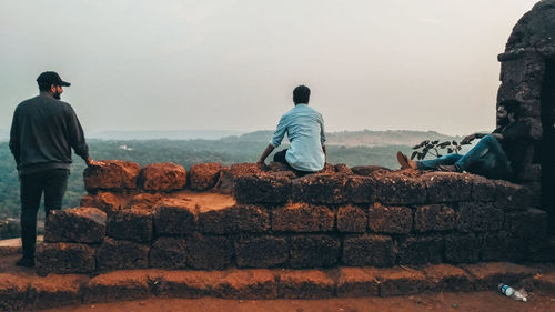 Rear view of men sitting on retaining wall against sky