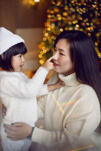 Beautiful korean mom and baby daughter sitting in white knitted sweaters at the christmas tree