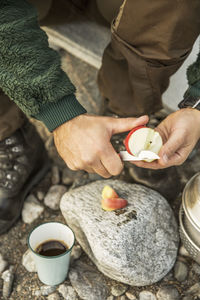 Midsection of man cutting apple over rock by coffee cup at camp