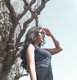 Low angle view of girl looking away while shielding eyes against tree trunk and sky