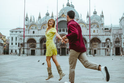 Happy couple running while holding hands in city against buildings and sky