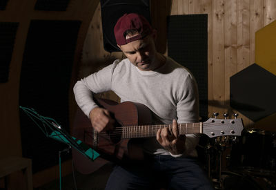 Caucasian man plays guitar,tuning musical instrument in hands, sitting in chair in studio. darken