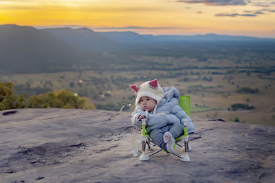 Full length of cute baby sitting on rock against landscape