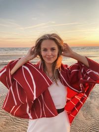 Portrait of young woman sitting at beach against sky during sunset