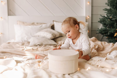 Funny funny little girl baby opens christmas gift box sitting on bed at home on holiday