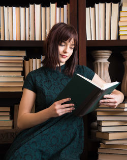 Young woman reading book while sitting against bookshelf in library