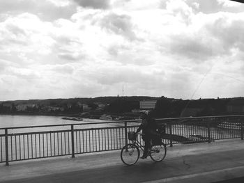 Man riding bicycle on bridge over river against sky