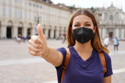 Optimistic traveler woman wearing black protective mask kn95 ffp2 showing thumbs up in city square