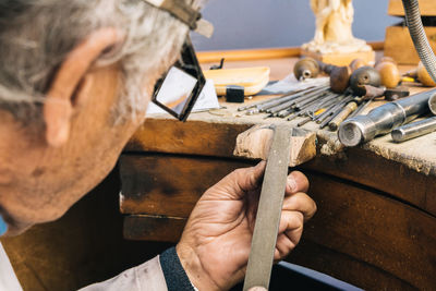 Side view of crop skilled mature craftsman in special glasses using polishing hand tool while creating jewelry accessory in workshop