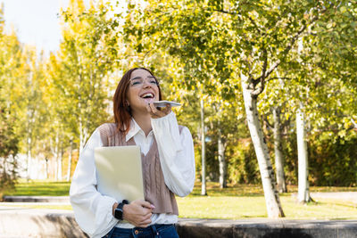 Positive smiling woman with glasses recording audio message while holding laptop in the park.