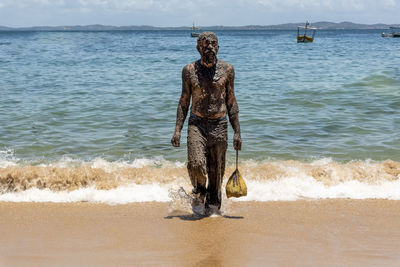 Man covered in oil walking on the sandy beach with the sea in the background.