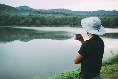 Side view of man holding mug while standing at lakeshore